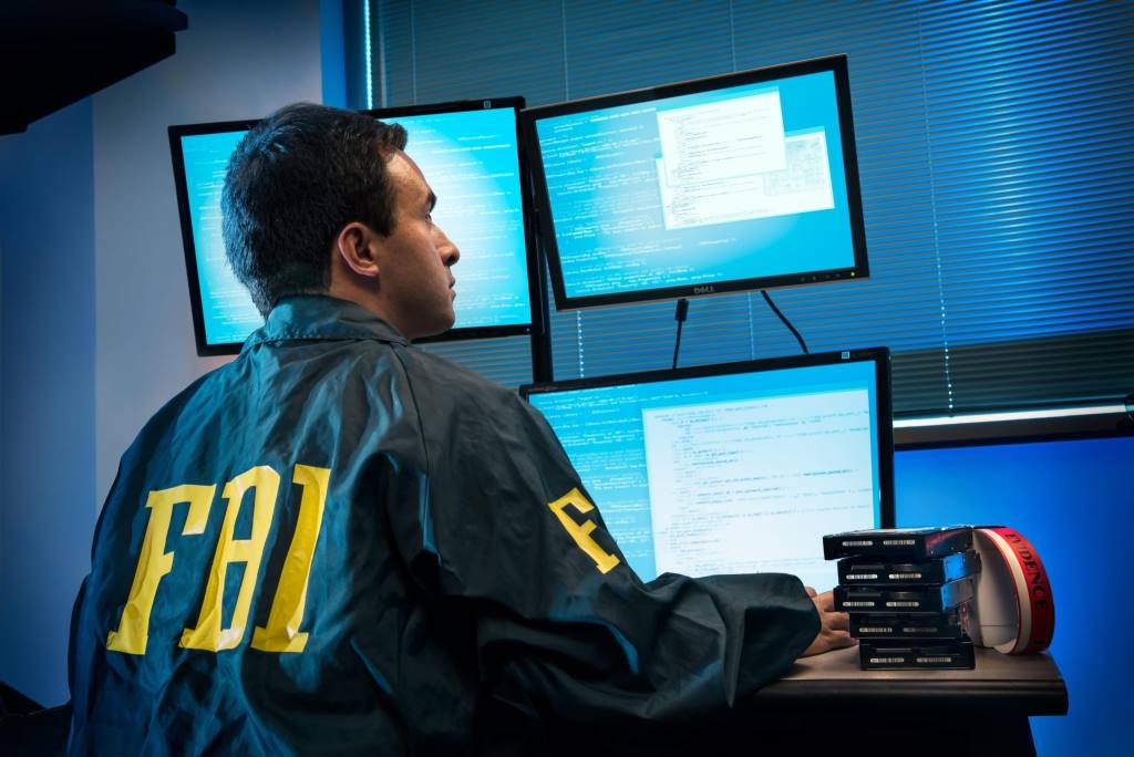 A man wearing an FBI jacket looking at multiple computer screens